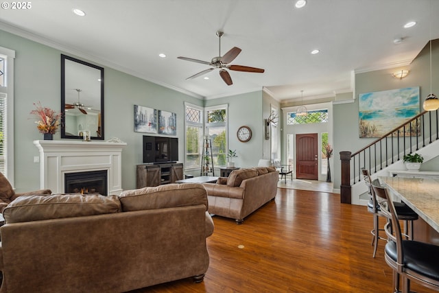 living room featuring a ceiling fan, wood finished floors, recessed lighting, ornamental molding, and a warm lit fireplace