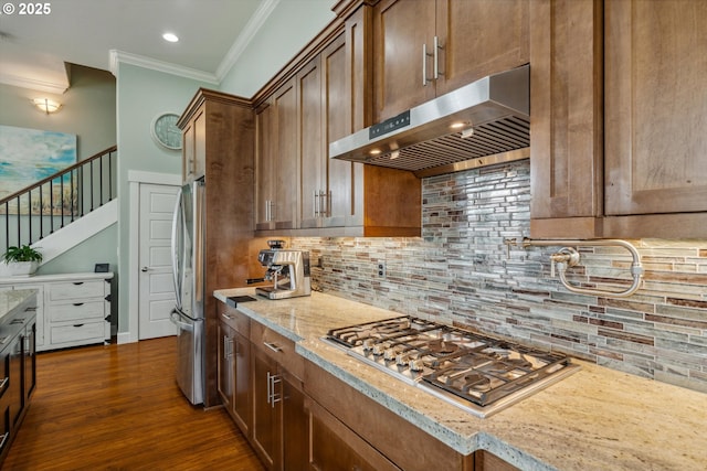 kitchen with under cabinet range hood, stainless steel appliances, ornamental molding, tasteful backsplash, and dark wood finished floors