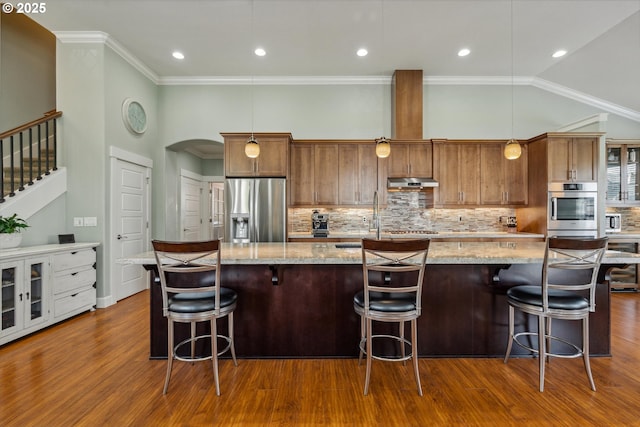 kitchen with appliances with stainless steel finishes, under cabinet range hood, dark wood-type flooring, a spacious island, and ornamental molding