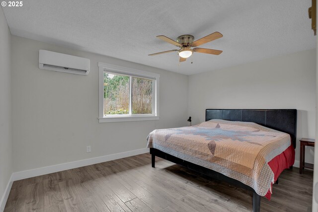 bedroom featuring an AC wall unit, ceiling fan, a textured ceiling, wood finished floors, and baseboards