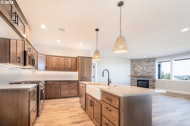 kitchen with an island with sink, sink, hanging light fixtures, stainless steel appliances, and light hardwood / wood-style flooring