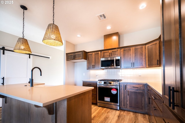 kitchen featuring hanging light fixtures, an island with sink, stainless steel appliances, a barn door, and backsplash