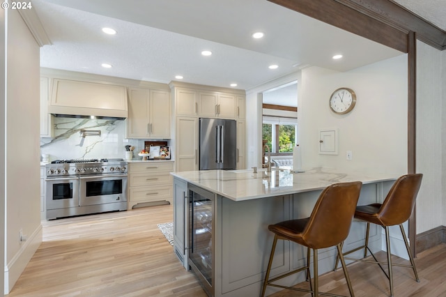 kitchen with wine cooler, light stone counters, stainless steel appliances, premium range hood, and a kitchen bar