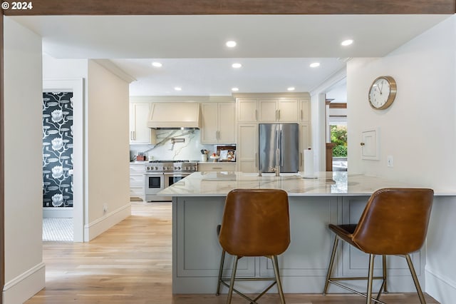 kitchen with a breakfast bar area, stainless steel appliances, premium range hood, light wood-type flooring, and light stone countertops