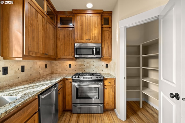 kitchen featuring brown cabinets, light wood-style flooring, glass insert cabinets, appliances with stainless steel finishes, and tasteful backsplash