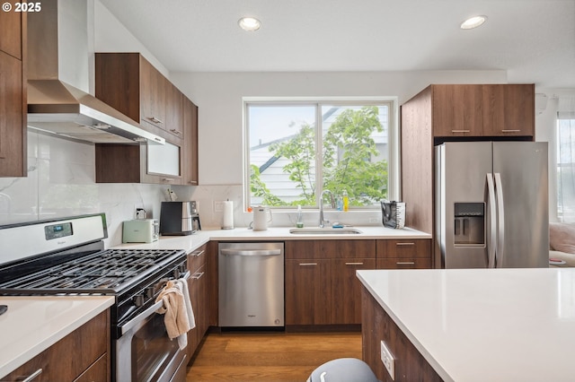 kitchen featuring wall chimney range hood, appliances with stainless steel finishes, a sink, light countertops, and light wood-style flooring