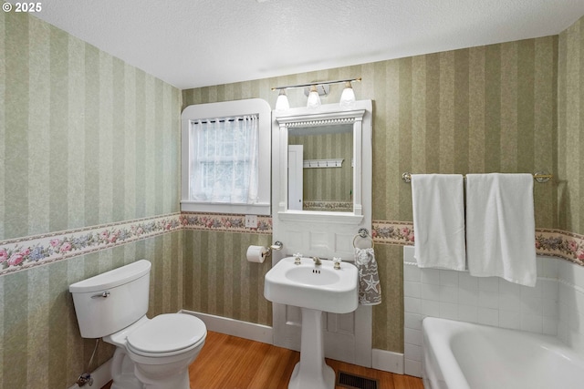full bath with toilet, wood finished floors, a textured ceiling, visible vents, and wallpapered walls