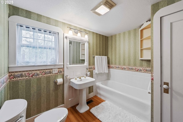 bathroom featuring wallpapered walls, toilet, a wainscoted wall, wood finished floors, and a textured ceiling