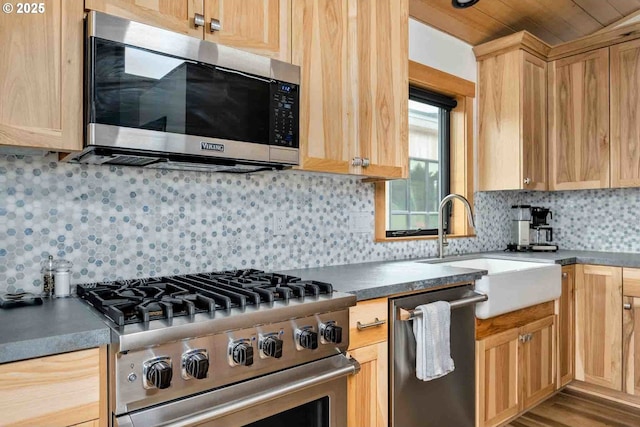 kitchen featuring appliances with stainless steel finishes, a sink, light brown cabinetry, decorative backsplash, and dark countertops