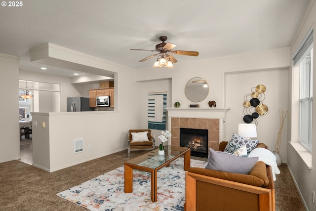 carpeted living area featuring ceiling fan with notable chandelier, a fireplace, visible vents, baseboards, and ornamental molding