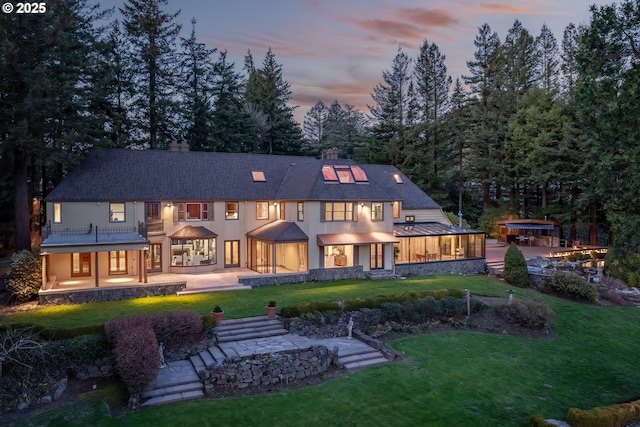 back of house at dusk with stucco siding, a lawn, a chimney, and a patio area