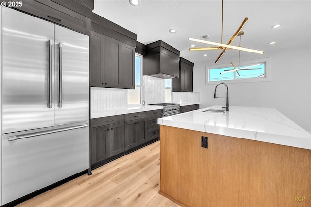 kitchen with decorative light fixtures, sink, stainless steel built in fridge, custom range hood, and light stone counters