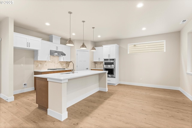 kitchen featuring light wood-type flooring, a kitchen island with sink, sink, decorative light fixtures, and white cabinetry