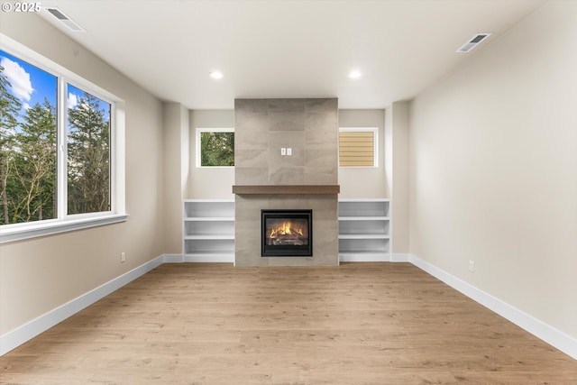 unfurnished living room featuring built in features, light hardwood / wood-style flooring, a healthy amount of sunlight, and a tiled fireplace