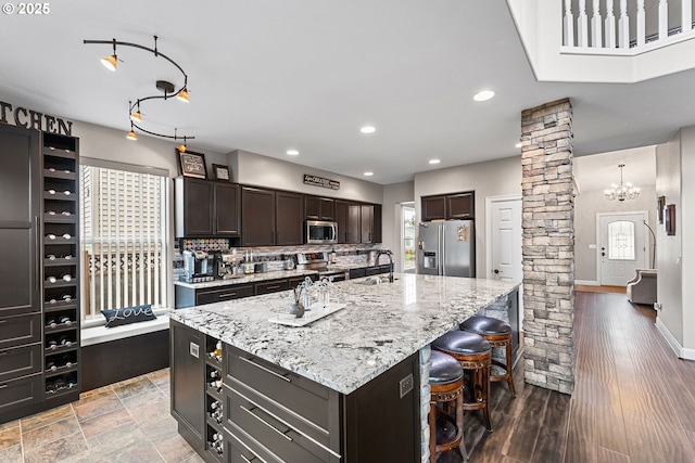 kitchen featuring ornate columns, appliances with stainless steel finishes, an island with sink, a breakfast bar area, and dark brown cabinetry