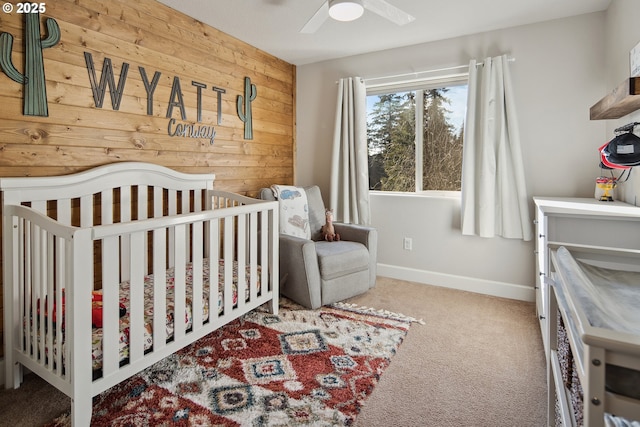 bedroom featuring wooden walls, a nursery area, ceiling fan, and carpet flooring