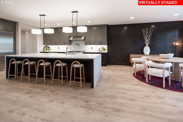 kitchen featuring a large island, light wood-type flooring, under cabinet range hood, a sink, and built in fridge