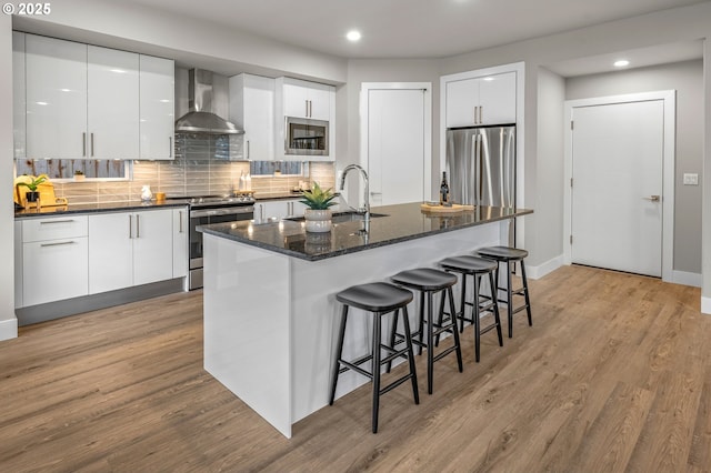 kitchen with a breakfast bar, light wood finished floors, decorative backsplash, appliances with stainless steel finishes, and wall chimney range hood