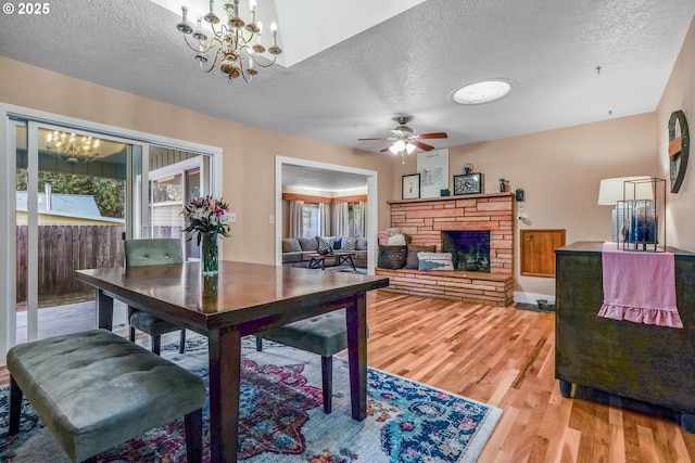 dining space featuring light wood-type flooring, a stone fireplace, a textured ceiling, and ceiling fan with notable chandelier