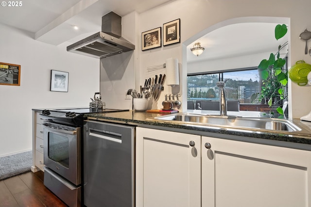 kitchen with dark wood-type flooring, stainless steel appliances, wall chimney range hood, white cabinetry, and a sink