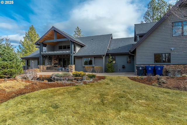 rear view of property with stone siding, roof with shingles, a patio, and a lawn