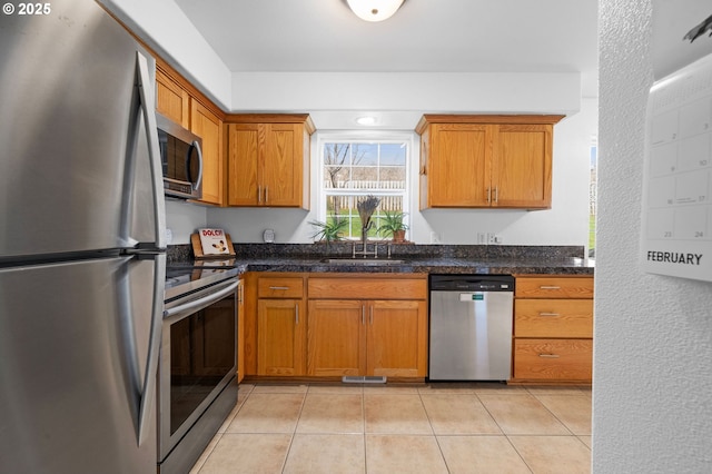 kitchen with brown cabinets, light tile patterned floors, stainless steel appliances, dark countertops, and a sink