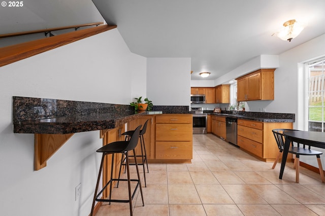 kitchen with stainless steel appliances, brown cabinetry, light tile patterned flooring, a peninsula, and a kitchen bar