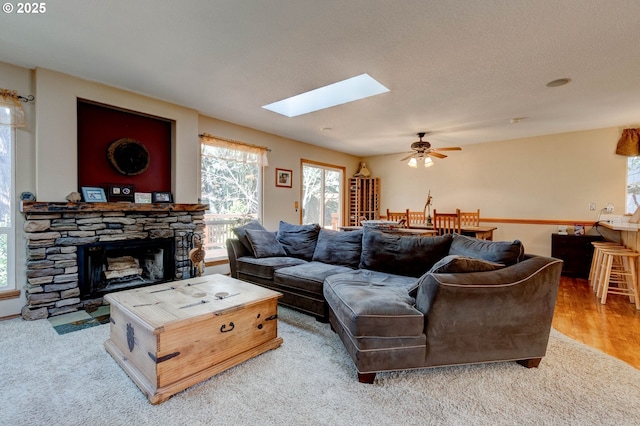 living room featuring ceiling fan, light wood-type flooring, a stone fireplace, and a skylight