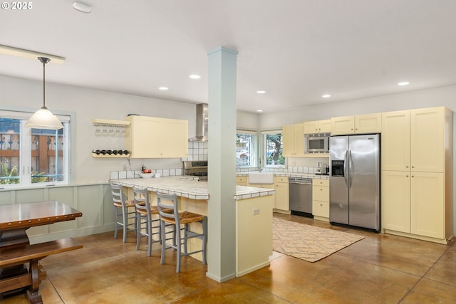 kitchen with wall chimney range hood, tile counters, a peninsula, stainless steel appliances, and a sink