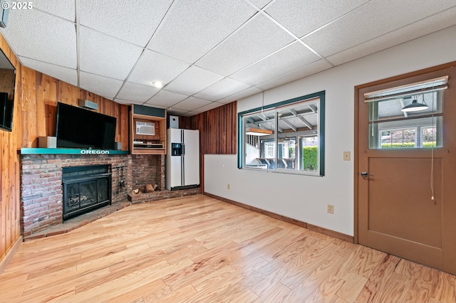unfurnished living room featuring light wood-style floors, a brick fireplace, wood walls, baseboards, and a drop ceiling