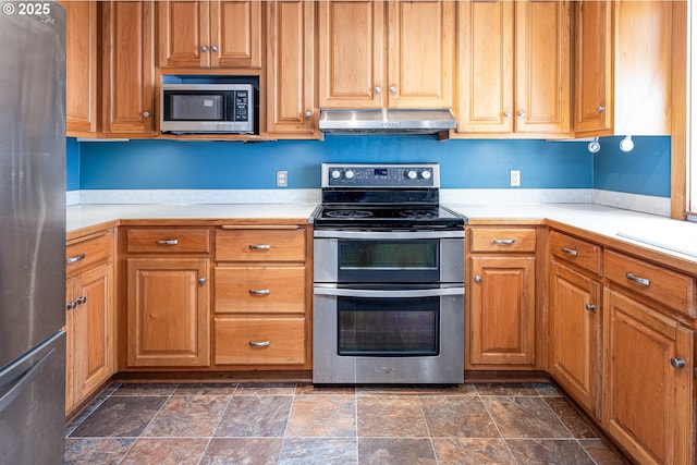 kitchen with under cabinet range hood, light countertops, brown cabinets, stone finish floor, and stainless steel appliances