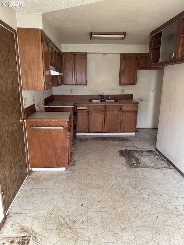 kitchen with baseboards, under cabinet range hood, brown cabinetry, stone finish floor, and a sink