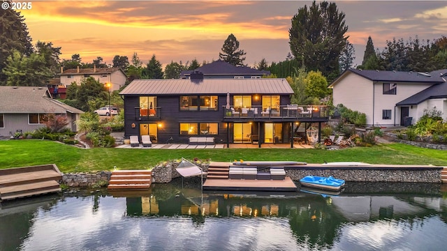 back house at dusk with a deck with water view, a patio, a balcony, and a lawn