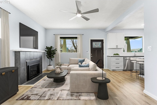 living area with a ceiling fan, baseboards, light wood-style floors, beam ceiling, and a glass covered fireplace