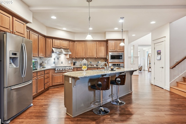 kitchen featuring a kitchen bar, hanging light fixtures, appliances with stainless steel finishes, an island with sink, and light stone countertops