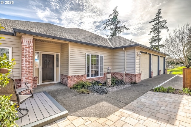 view of exterior entry with a garage, brick siding, driveway, and a shingled roof