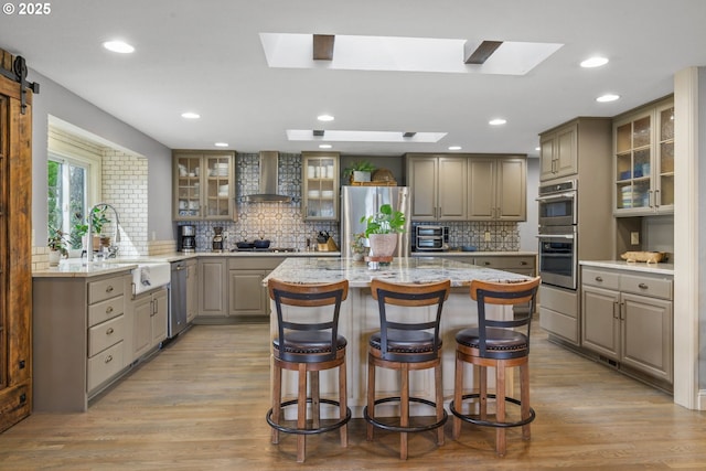 kitchen with wall chimney range hood, stainless steel appliances, gray cabinetry, a sink, and a kitchen breakfast bar