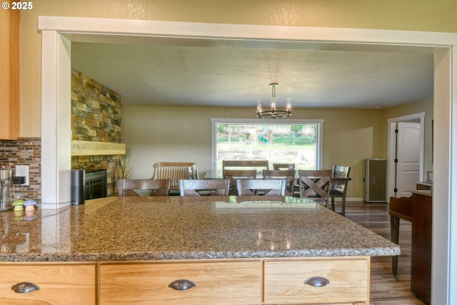 kitchen with backsplash, wood finished floors, dark stone counters, a stone fireplace, and a chandelier