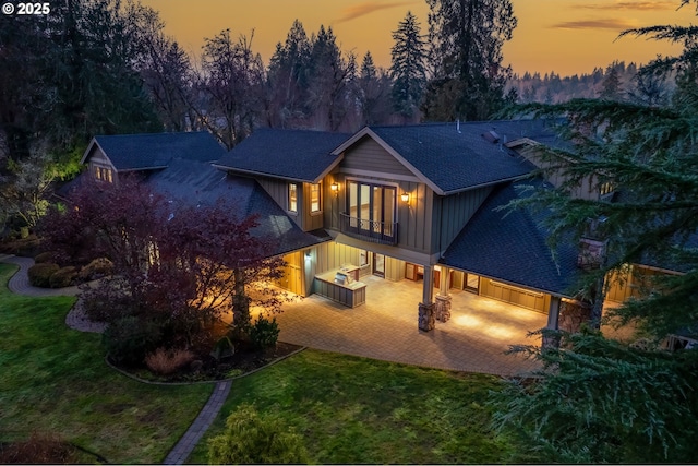 back house at dusk with a balcony, a patio, and a lawn