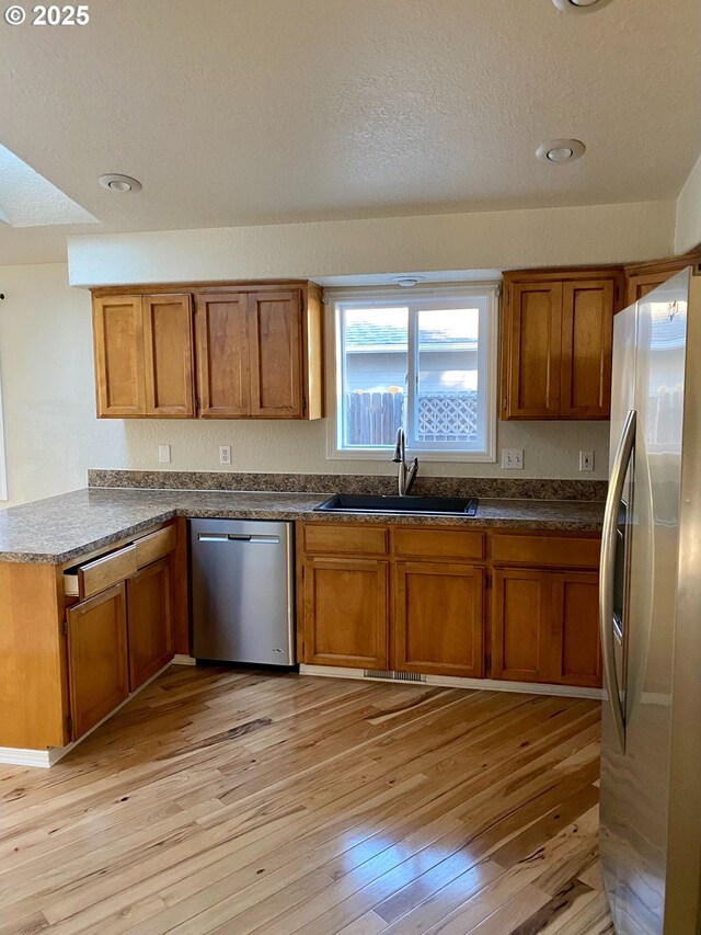 kitchen with appliances with stainless steel finishes, a sink, a textured ceiling, brown cabinetry, and light wood-type flooring
