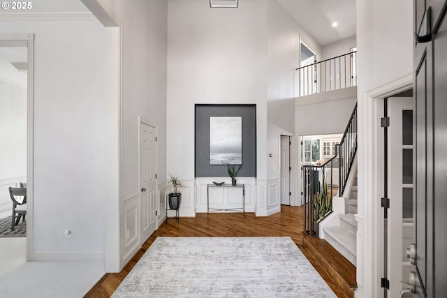 foyer with wood finished floors, stairs, a towering ceiling, a wainscoted wall, and crown molding