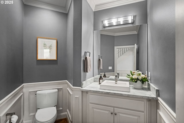 half bathroom with a decorative wall, wainscoting, toilet, crown molding, and vanity