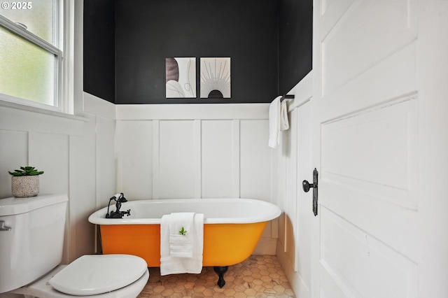 bathroom featuring tile patterned flooring, a decorative wall, toilet, a wainscoted wall, and a soaking tub