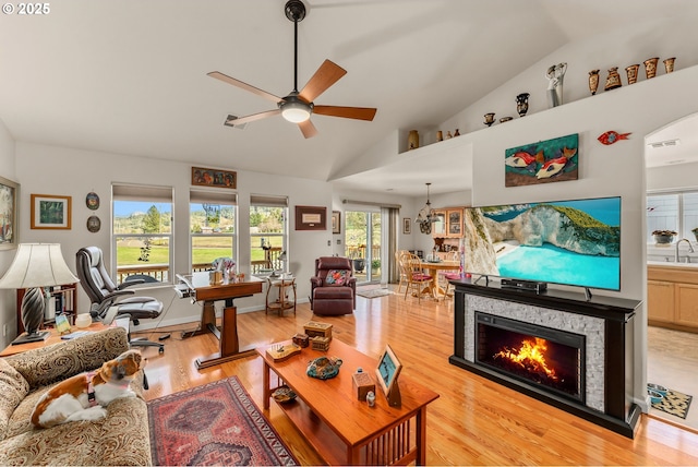 living room featuring ceiling fan, high vaulted ceiling, a stone fireplace, baseboards, and light wood-type flooring