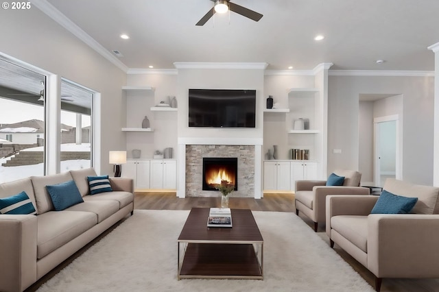 living room with light hardwood / wood-style floors, ornamental molding, built in features, ceiling fan, and a stone fireplace