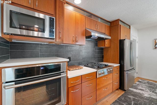 9004 SW 36th Ave, Portland, OR 97219 - kitchen featuring stainless steel appliances, under cabinet range hood, brown cabinetry, light wood-style flooring, and a textured ceiling