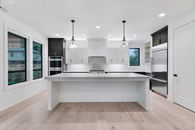 kitchen with stainless steel appliances, light wood-style floors, light countertops, an island with sink, and custom range hood