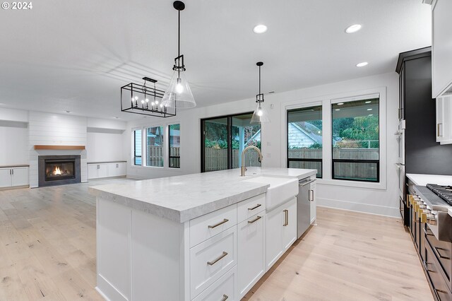 kitchen with a large fireplace, light wood finished floors, decorative light fixtures, a kitchen island with sink, and stainless steel dishwasher