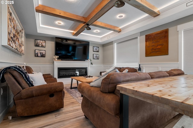 home theater featuring a decorative wall, beamed ceiling, light wood-style flooring, coffered ceiling, and a glass covered fireplace