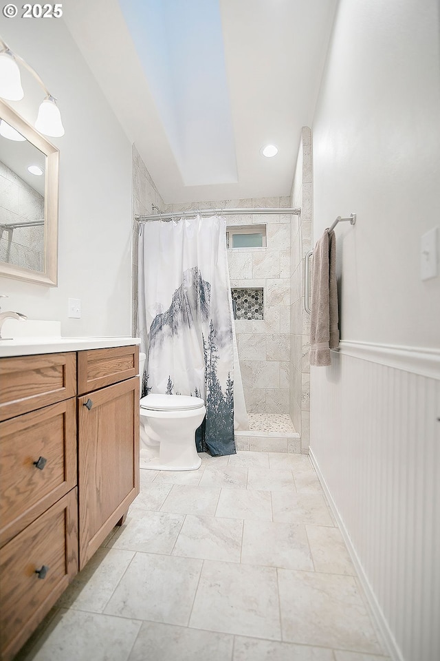 full bathroom with toilet, tiled shower, vanity, and wainscoting